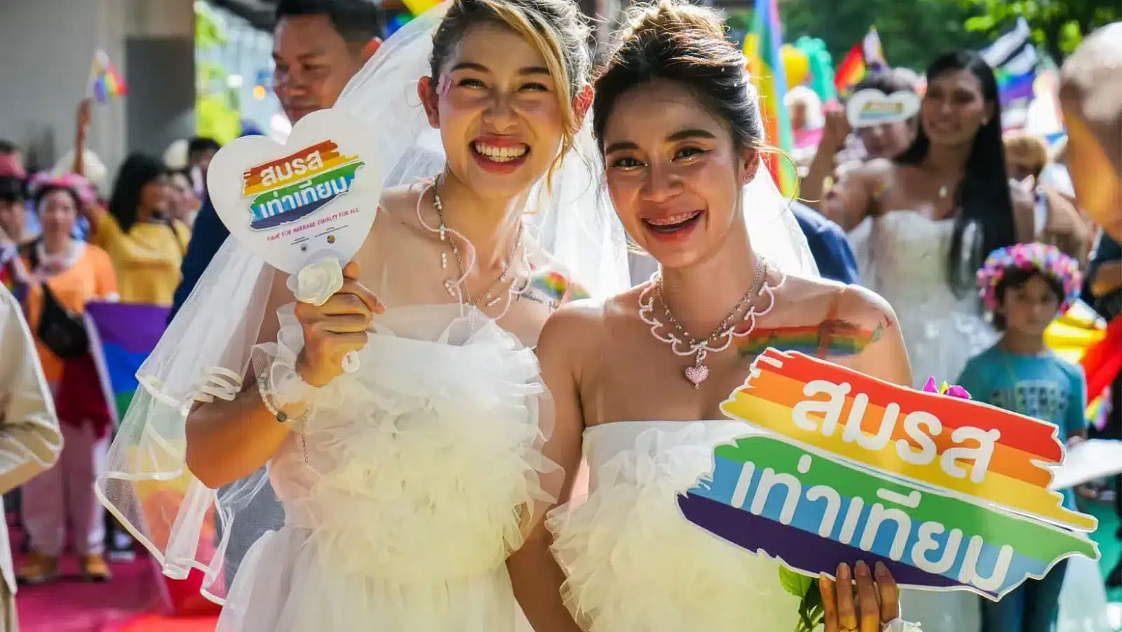 Two brides at a marriage equality celebration in Bangkok, surrounded by a crowd with rainbow flags