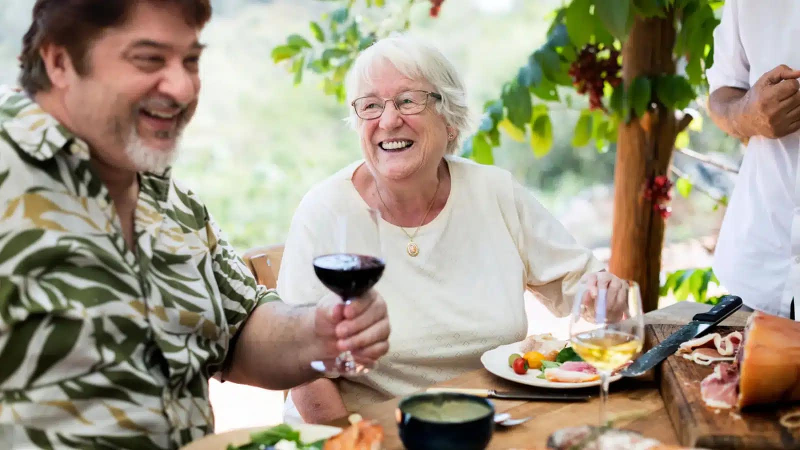 Two older people laughing over lunch and a glass of wine under a grapevine