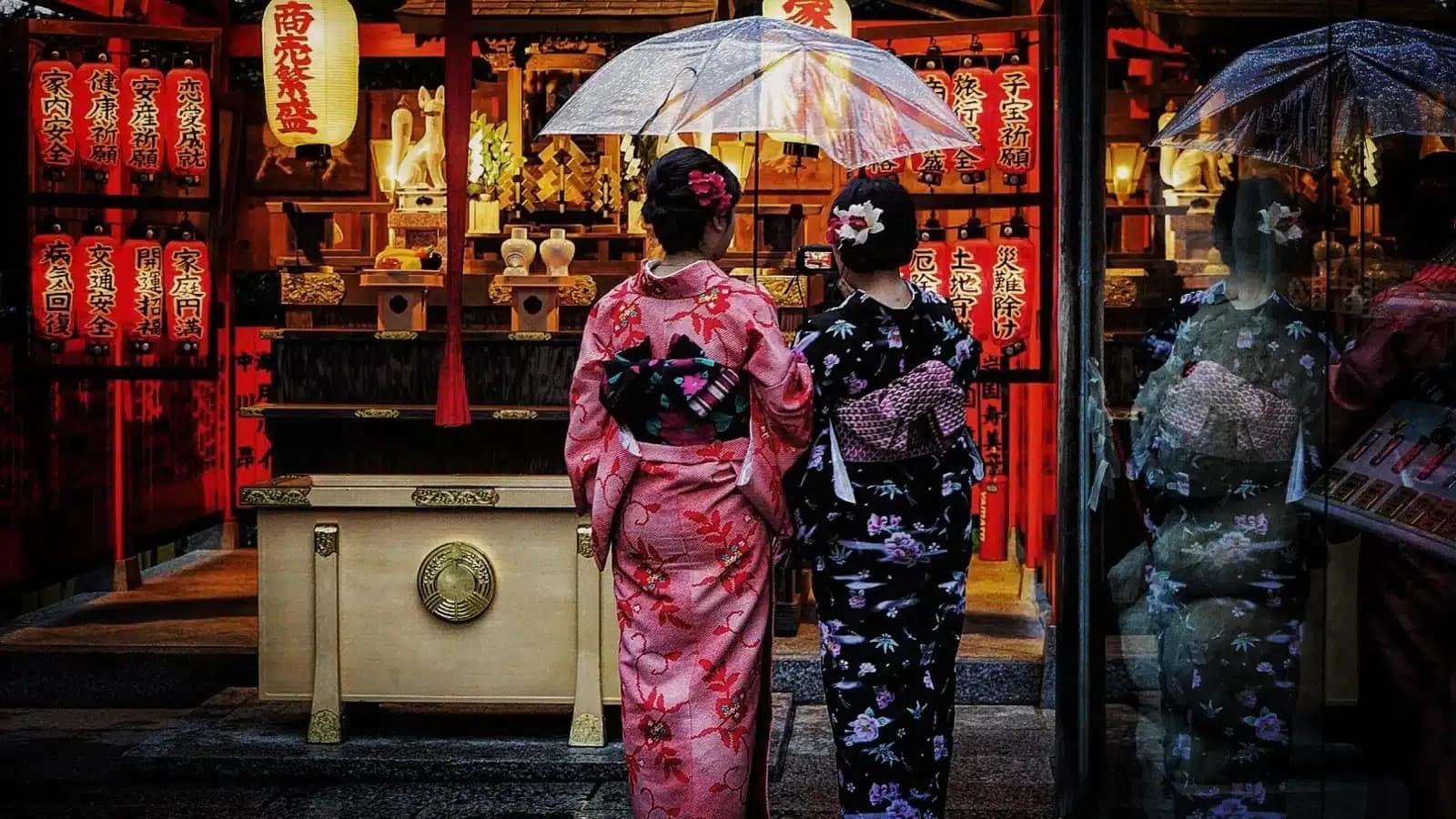 Two women in kimono at a shrine offering box hung with red paper lanterns in Japan